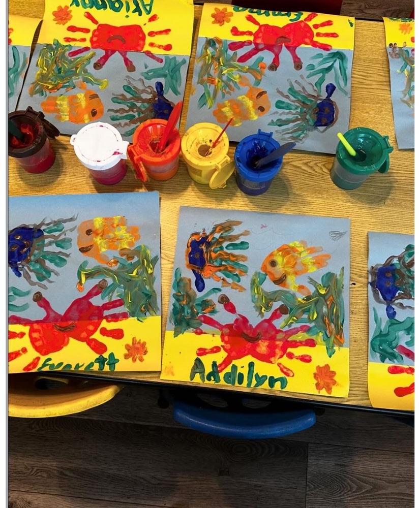 Handprint and fingerprint paintings of sea creatures drying on an activity table between a row of lidded paint cups