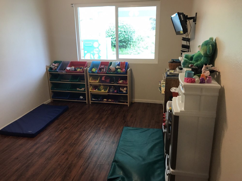 The indoor playroom at Gemma's home, with nap mats on the floor, toy bins under the window, a play kitchen and a shelf of books